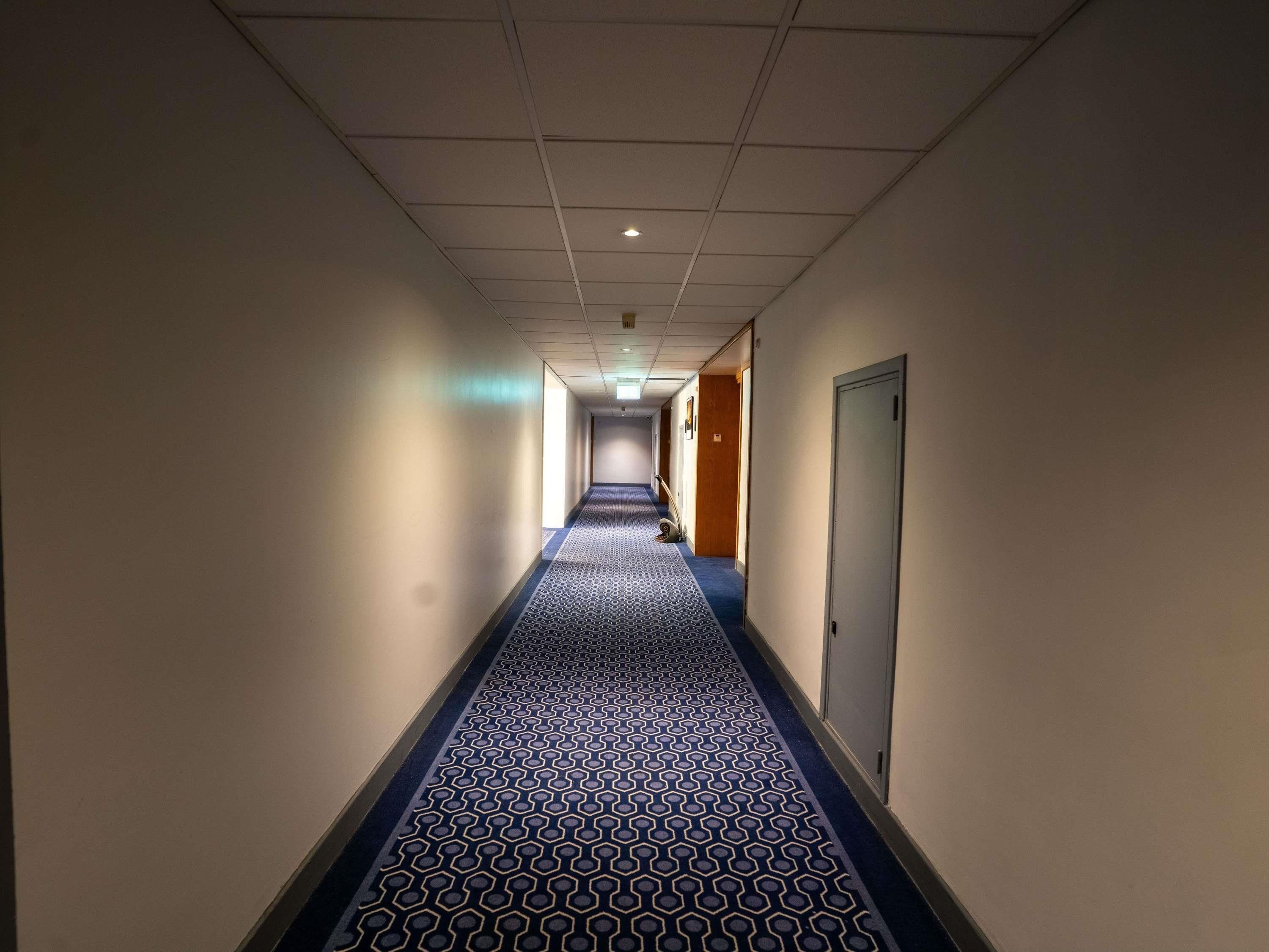 A hotel hallway with a patterned carpet, doors on both sides, and a ceiling with recessed lighting.