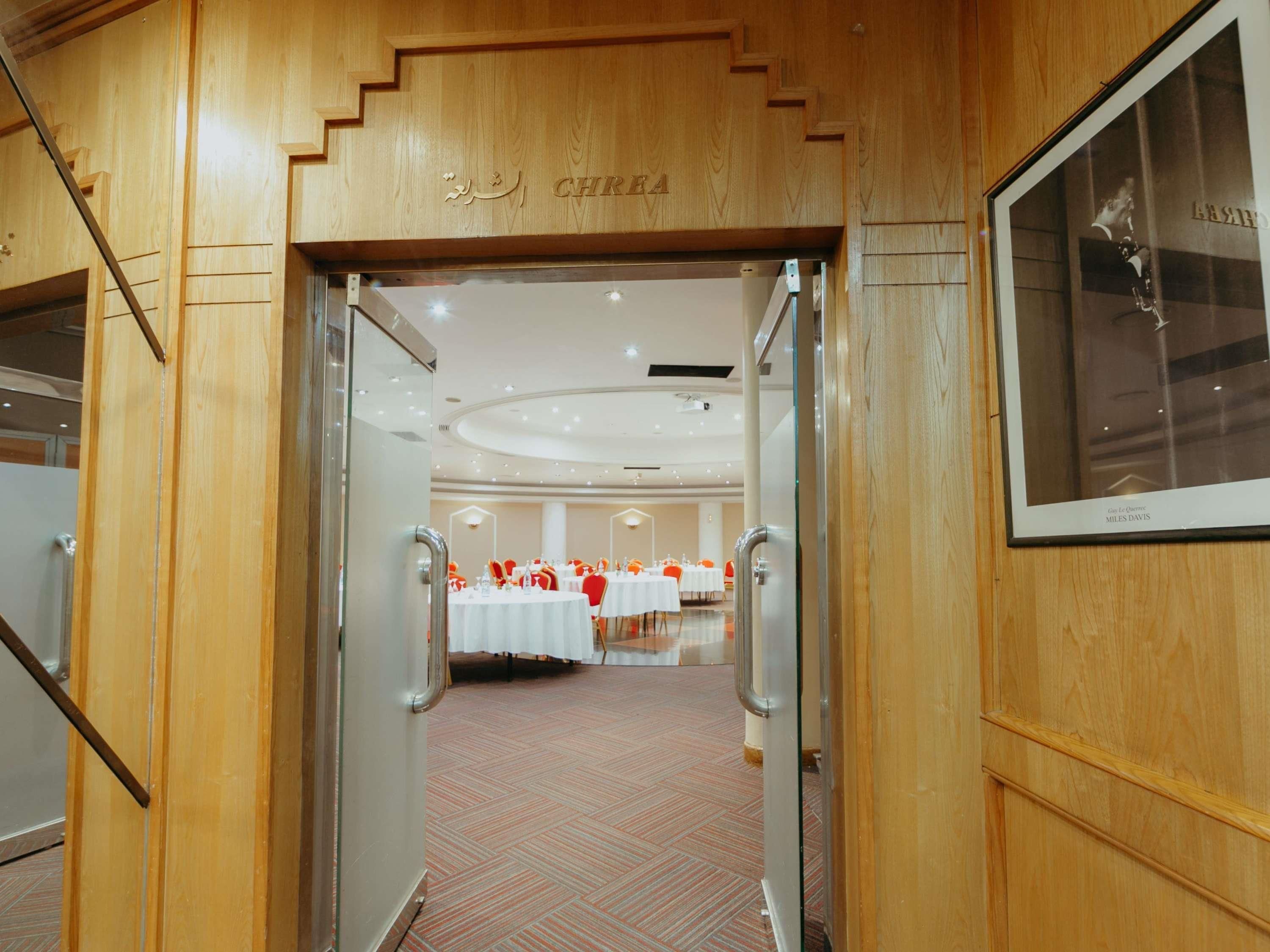 Entrada de un hotel con fachada de madera, un cuadro enmarcado en la pared y vistas a un salón de banquetes con mesas y sillas.