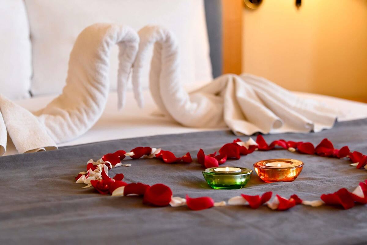 A bed with rose petals and candles, a white pillow, and a wall-mounted light fixture.