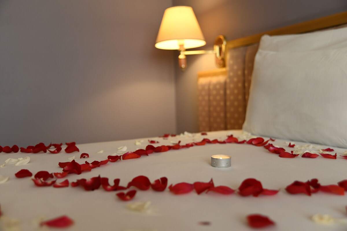 A bed with rose petals and a lit candle, a lamp, and a headboard.