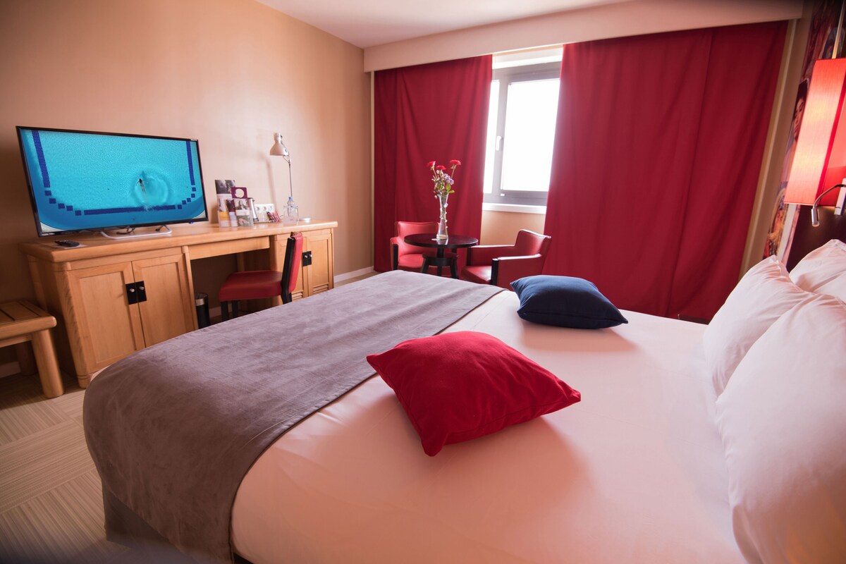 A hotel room with a bed, a desk with a computer, a chair, a small table with a vase, and a window with red curtains.