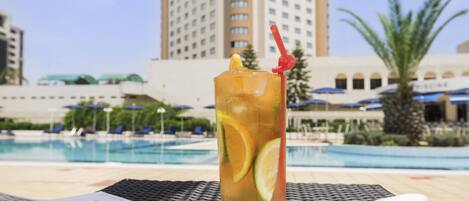 Un vaso alto con pajita y una rodaja de limón sobre una mesa junto a una piscina, con una revista y una toalla al lado.