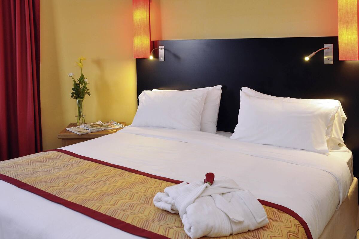 A hotel room with a neatly made bed, a bedside table with a vase of flowers, and a wall-mounted light fixture.
