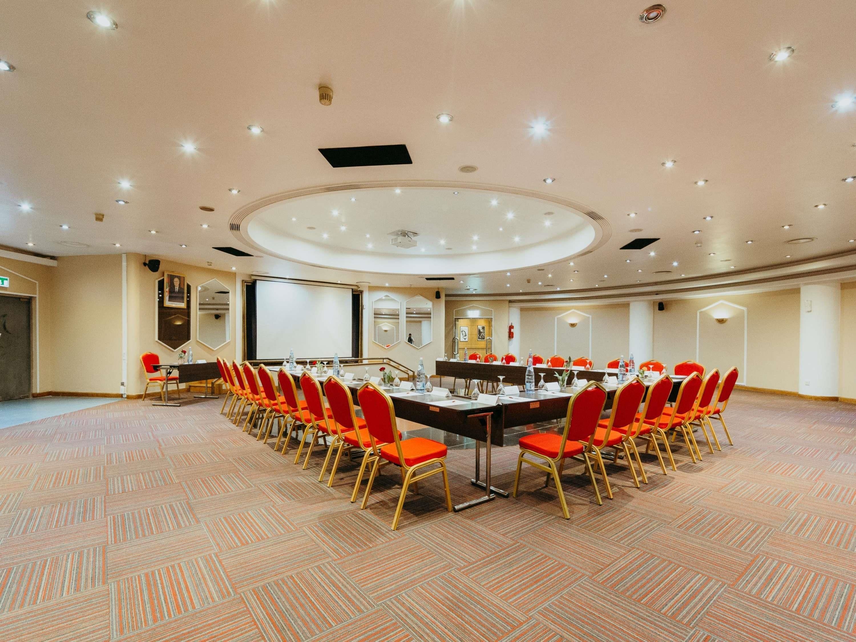 Un salón de conferencias con una larga mesa, sillas rojas y una pantalla de proyector.