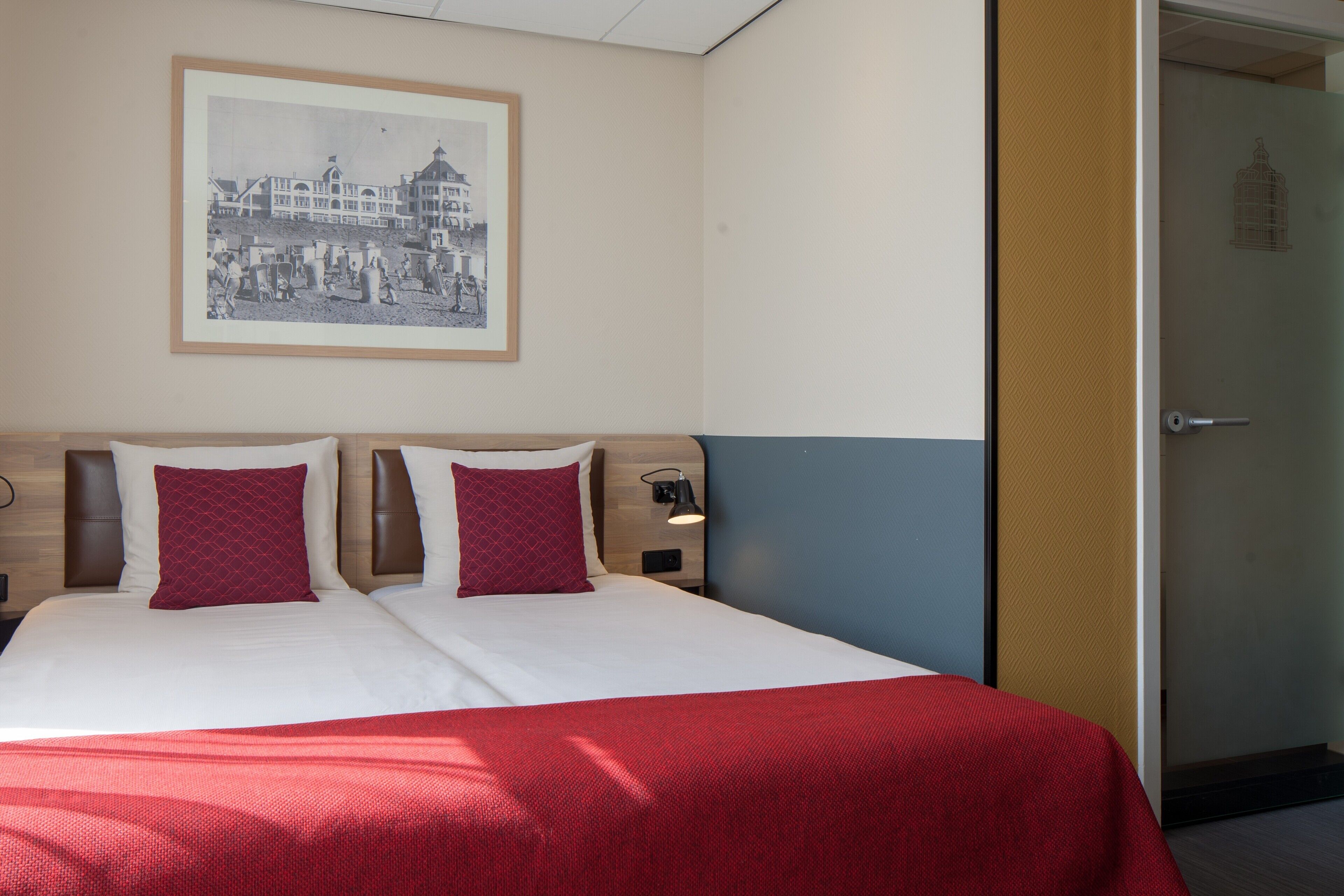 A double bed with a red blanket, two red pillows, and a framed picture on the wall.