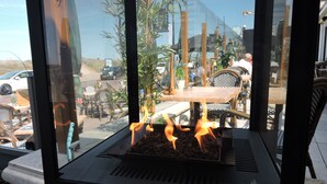 A grill with flames, surrounded by outdoor furniture and plants, with a view of a parking area and a building in the background. - Two Brothers Noordwijk Beach (Noordwijk)