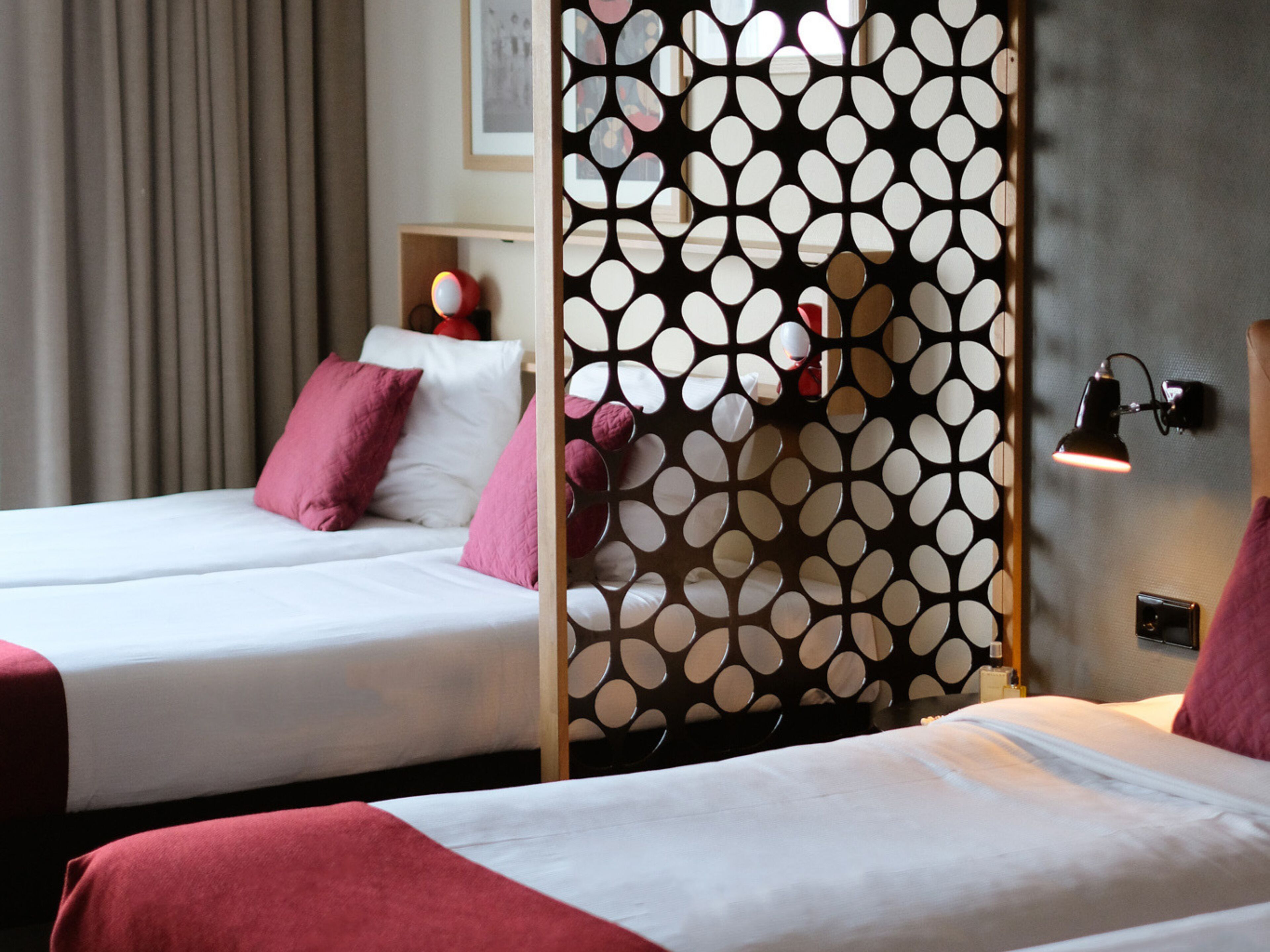 A hotel room with two beds, each with white bedding and pink pillows. There is a decorative screen with circular cutouts between the beds. A small table with a red lamp and a framed picture is visible in the background.