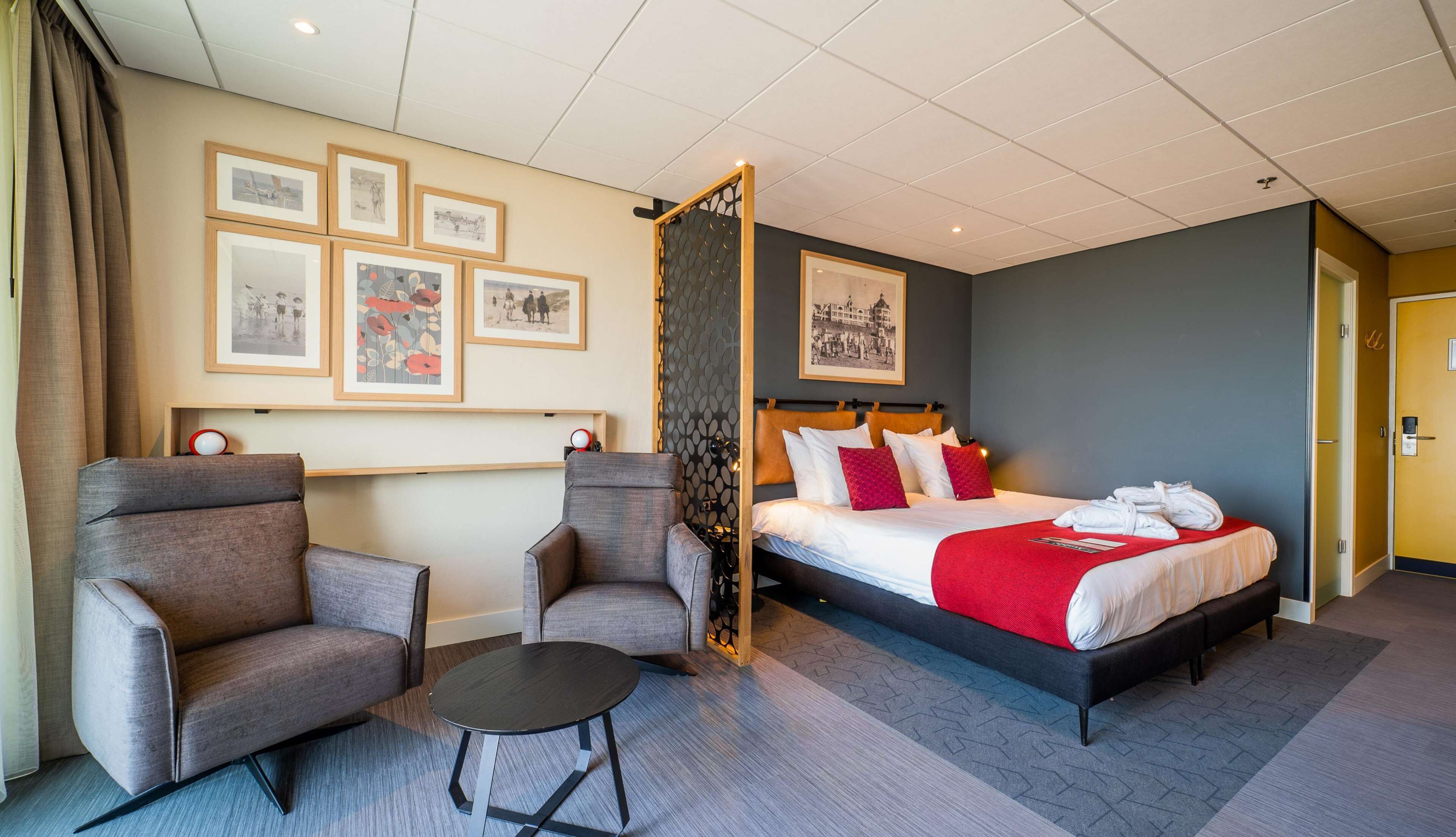 Et hotelværelse med en seng, to lænestole, et lille bord og billeder i rammer på væggen.