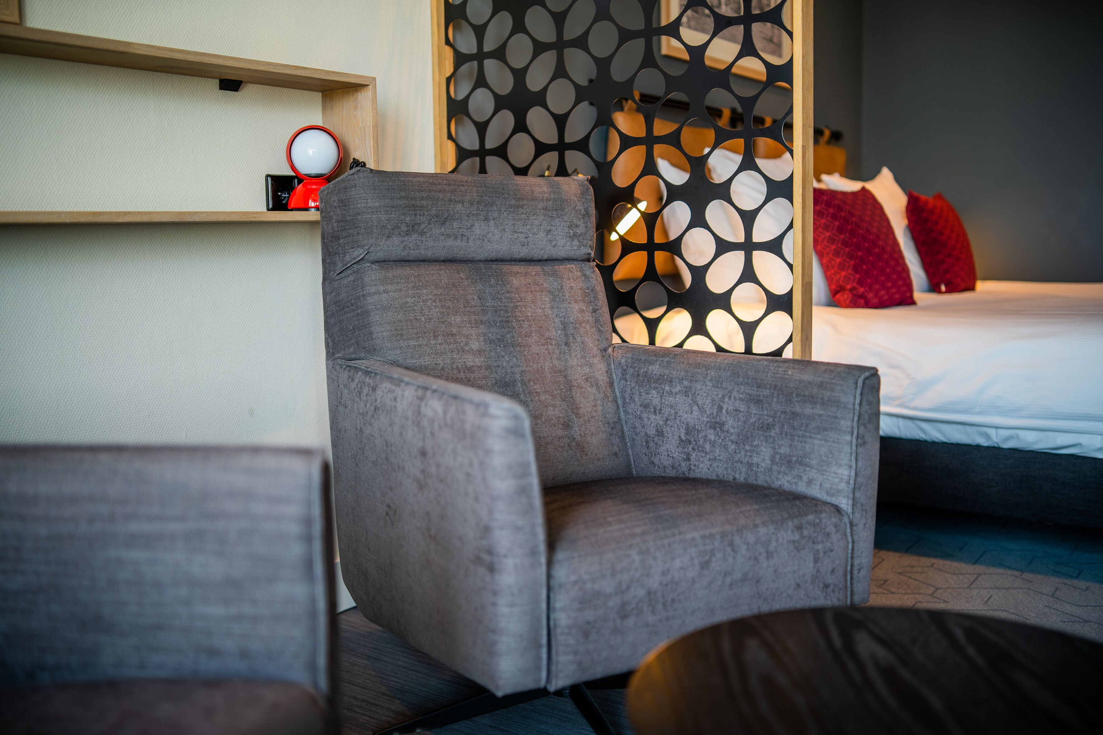 Une chambre d’hôtel moderne avec un fauteuil gris, une étagère en bois sur laquelle se trouve un objet rouge, et un panneau décoratif à motifs.