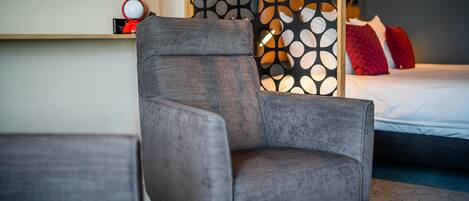 Une chambre d’hôtel moderne avec un fauteuil gris, une étagère en bois sur laquelle se trouve un objet rouge, et un panneau décoratif à motifs.