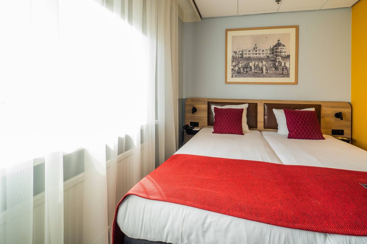 A hotel room with a bed, two pillows, a red blanket, a nightstand, a framed picture on the wall, and a window with curtains.