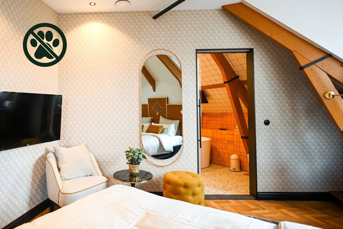 A bedroom with a bed, a TV, a mirror, a small table with a plant, a chair, and a door leading to a loft area.
