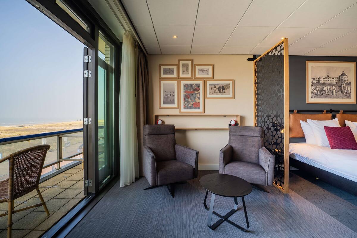 A hotel room with a balcony, two armchairs, a small table, a bed with pillows, and framed pictures on the wall.