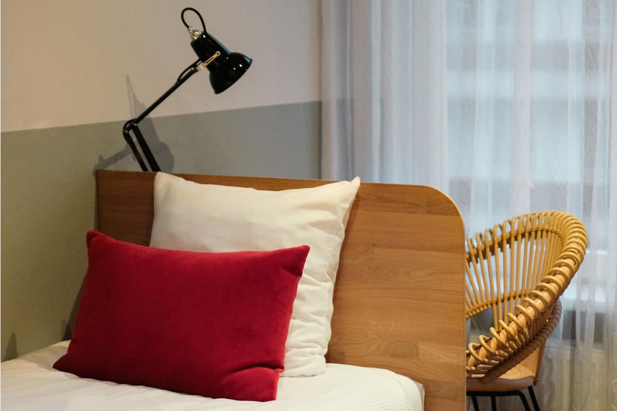 A bed with a red pillow, a white pillow, and a wooden headboard. There is a black desk lamp on the left side of the bed. A wicker chair is positioned to the right of the bed.