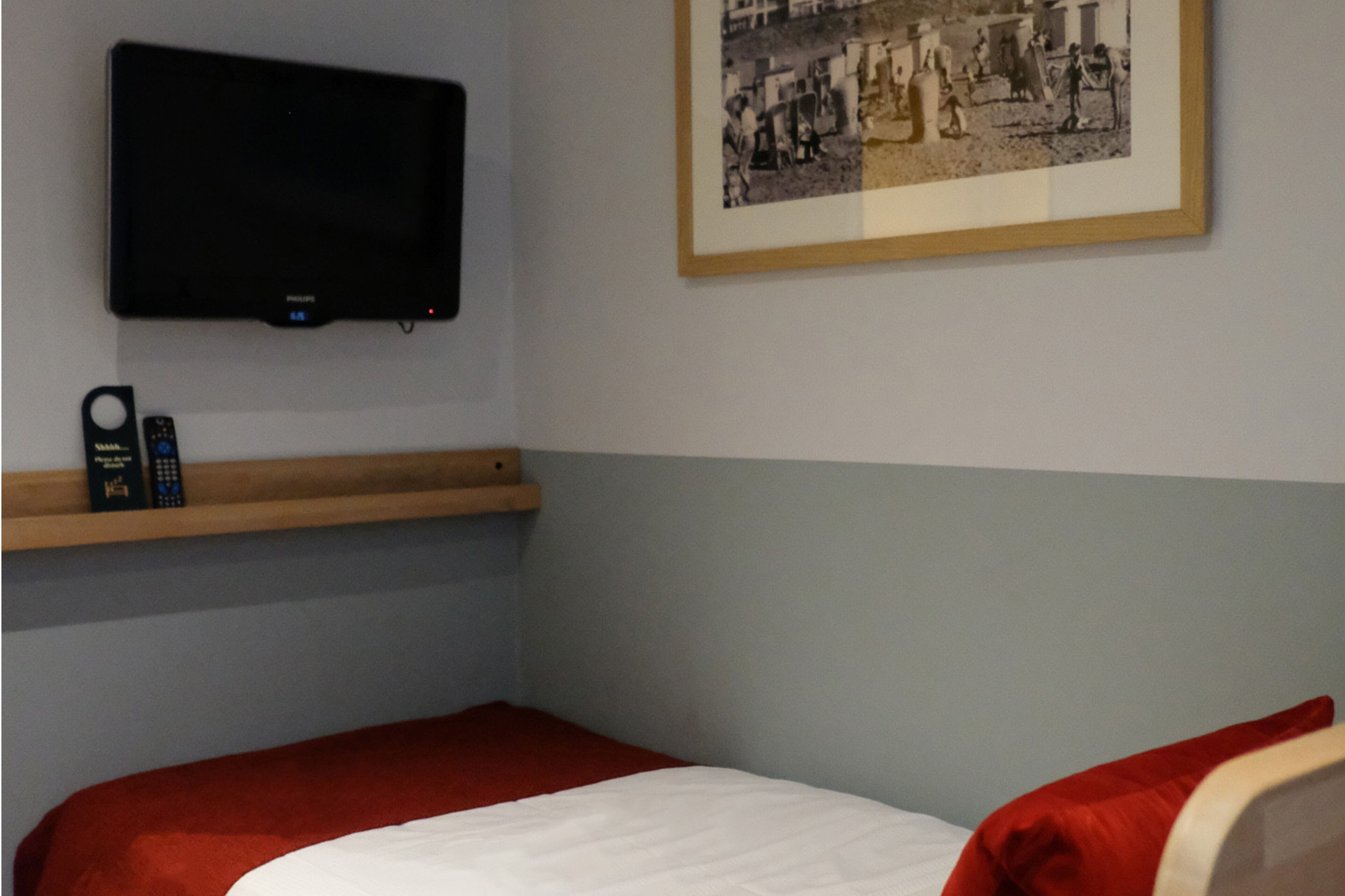 Un dormitorio con una cama, un televisor montado, un estante con un control remoto y un cuadro enmarcado en la pared.