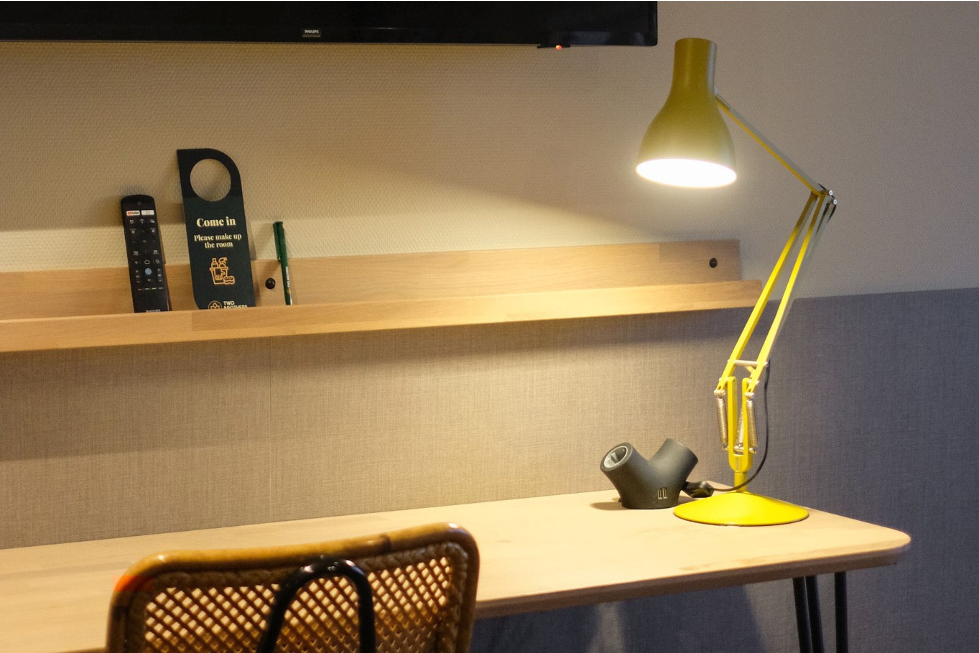 Un escritorio con una lámpara de escritorio amarilla, una silla y un estante con un letrero, controles remotos y un bolígrafo.