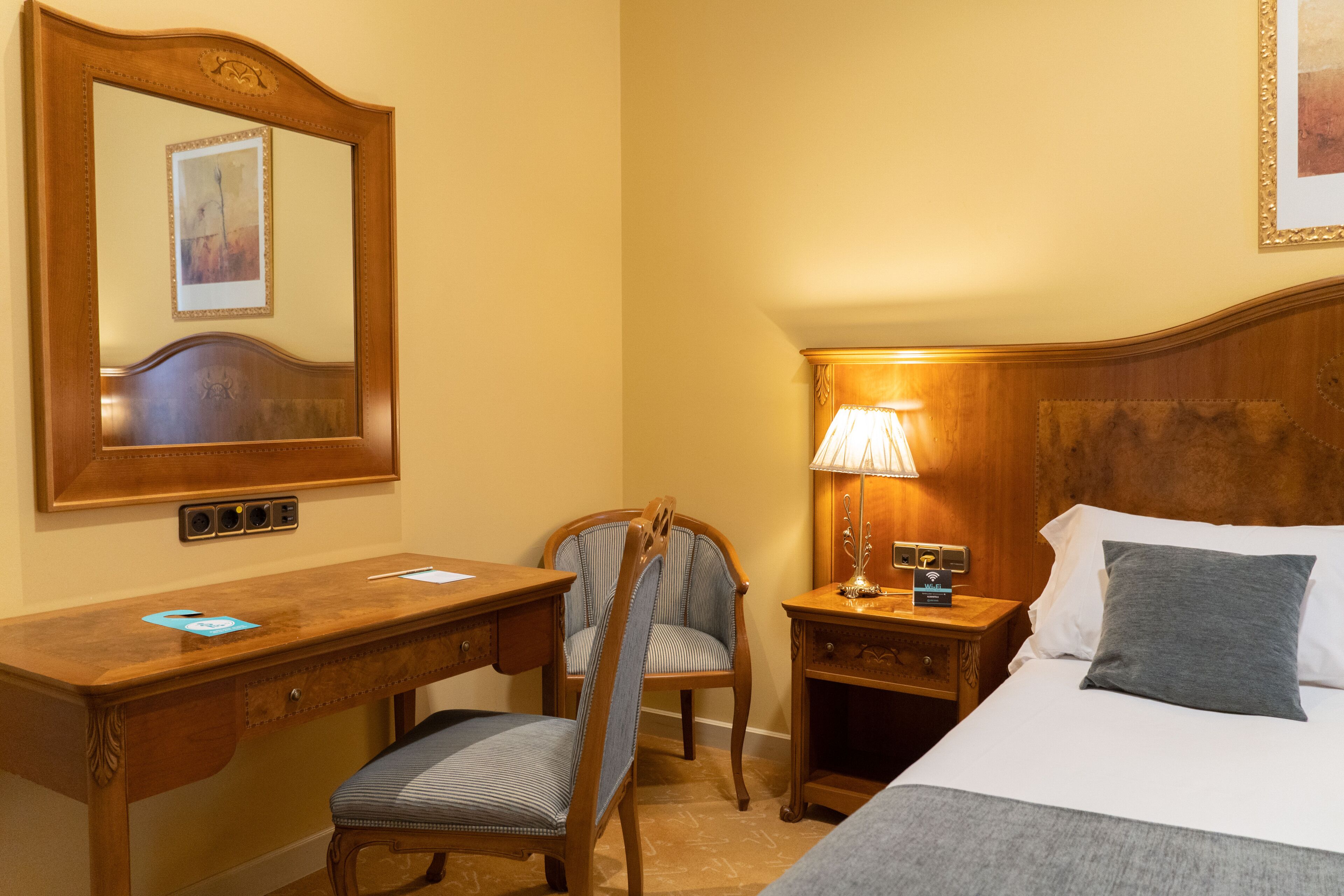 A hotel room with a wooden desk, a chair, a bedside table with a lamp, a bed with a pillow, and a large mirror on the wall.