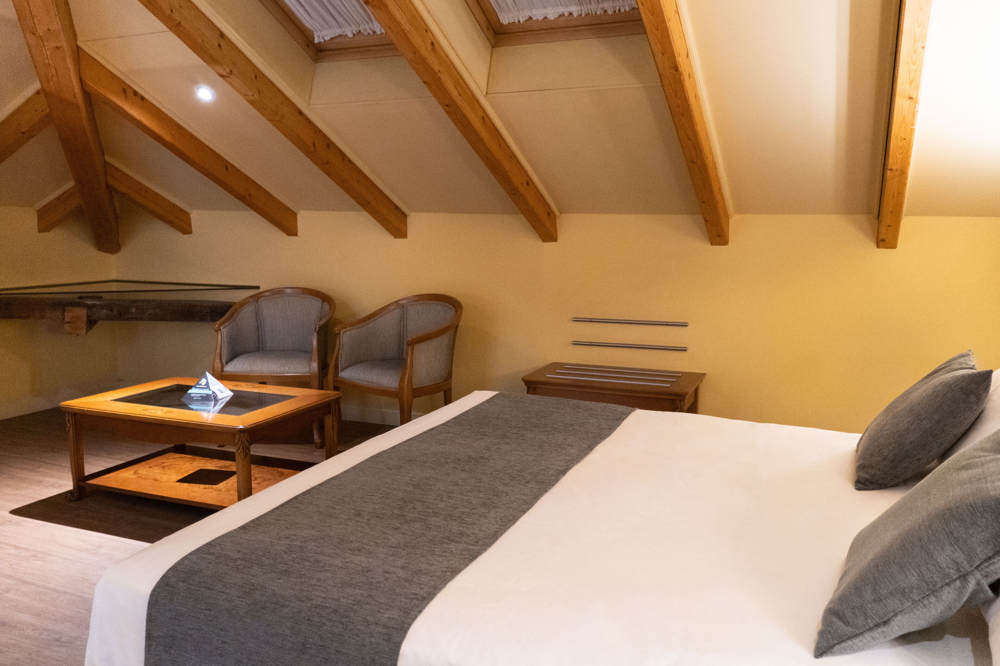 A bedroom with a bed, two chairs, a small table, wooden beams, and a sloped ceiling.
