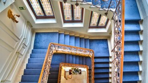 A grand staircase with ornate metal railings and blue carpeted steps, leading to a landing with a view of the sky through a glass ceiling.