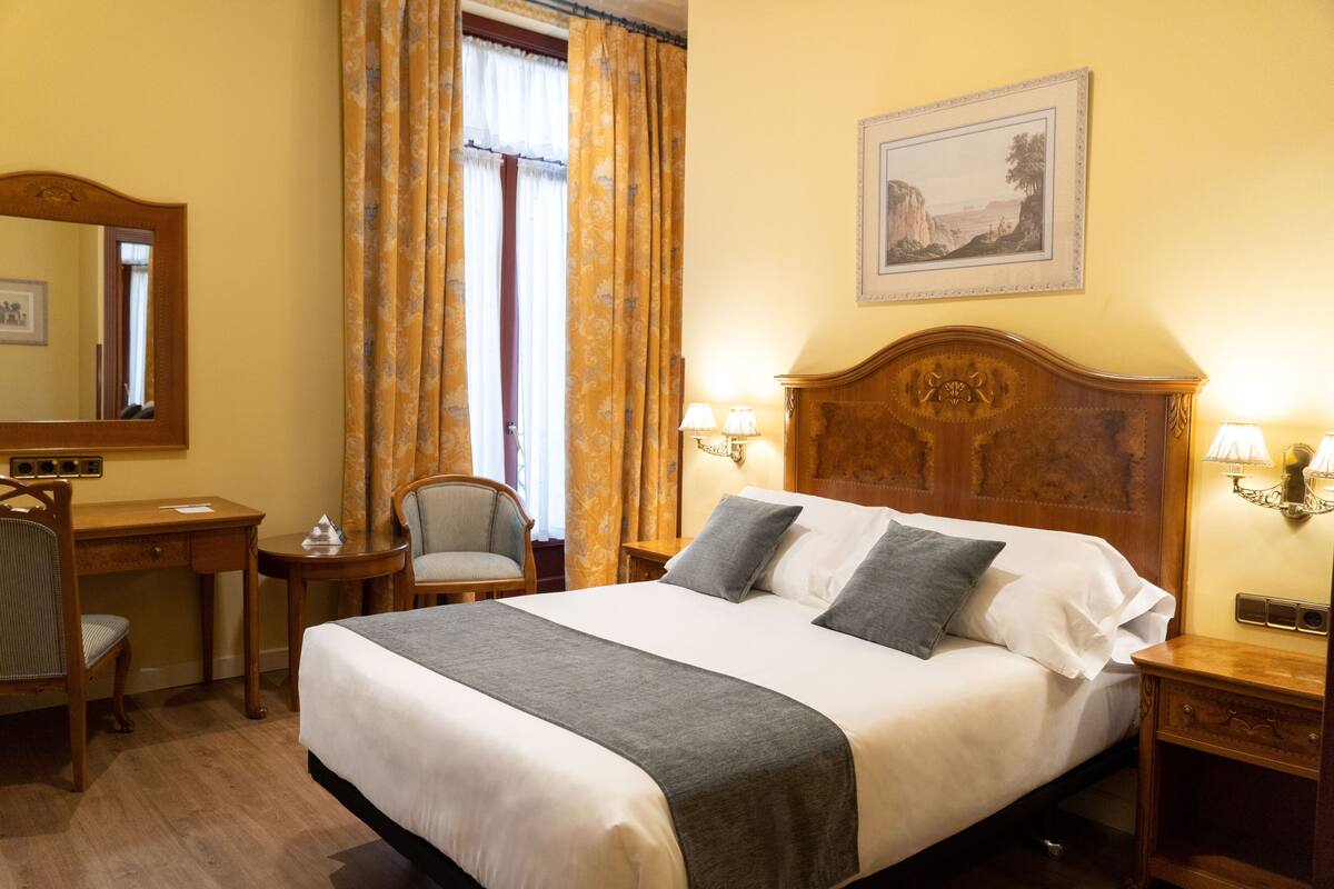 A hotel room with a large bed, a desk, a chair, and a nightstand with lamps.