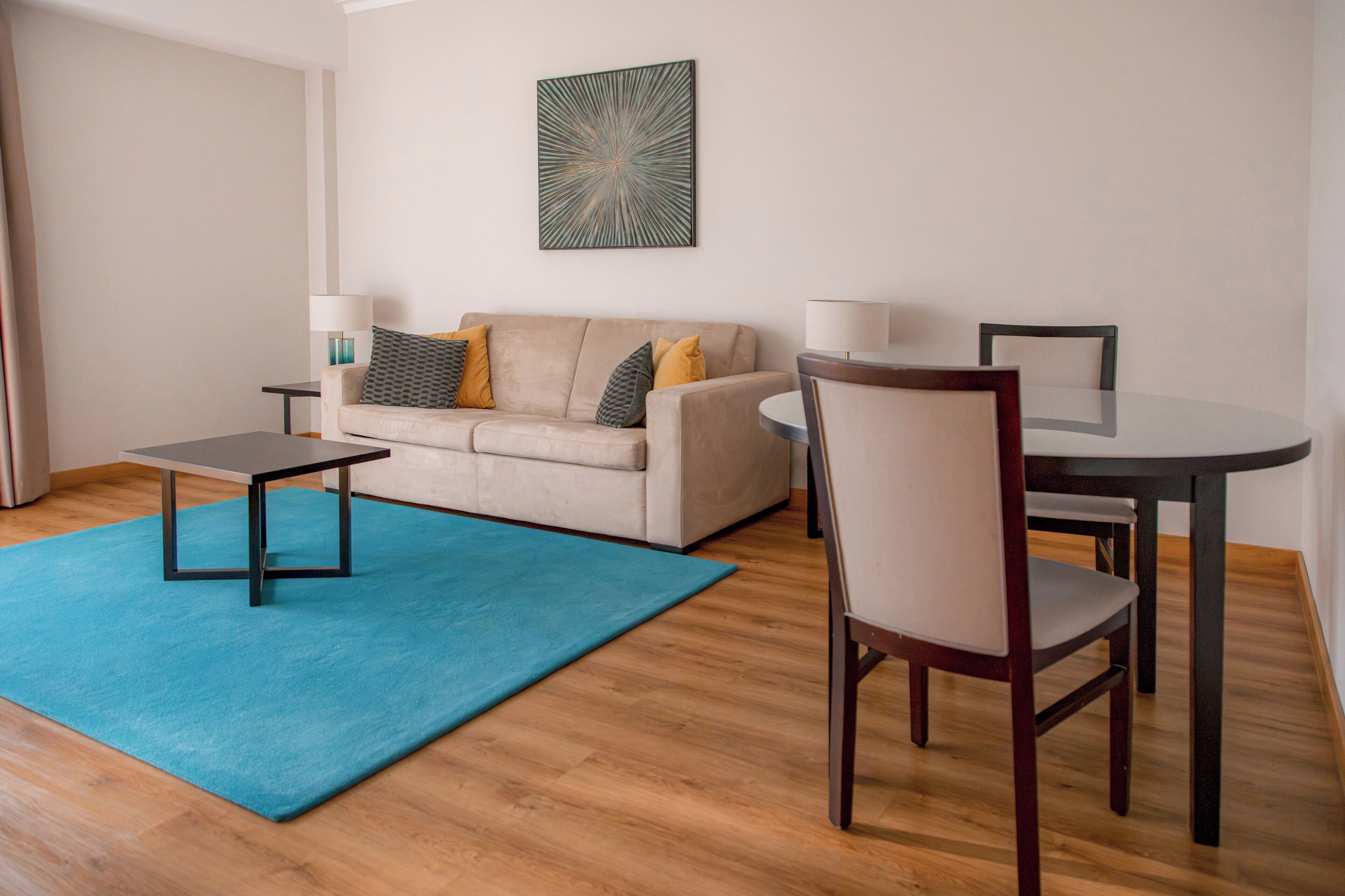A modern living room with a beige sofa, two side tables, a round dining table, and a blue rug.