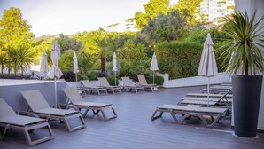 A rooftop lounge area with sun loungers and umbrellas, surrounded by greenery and a view of buildings.