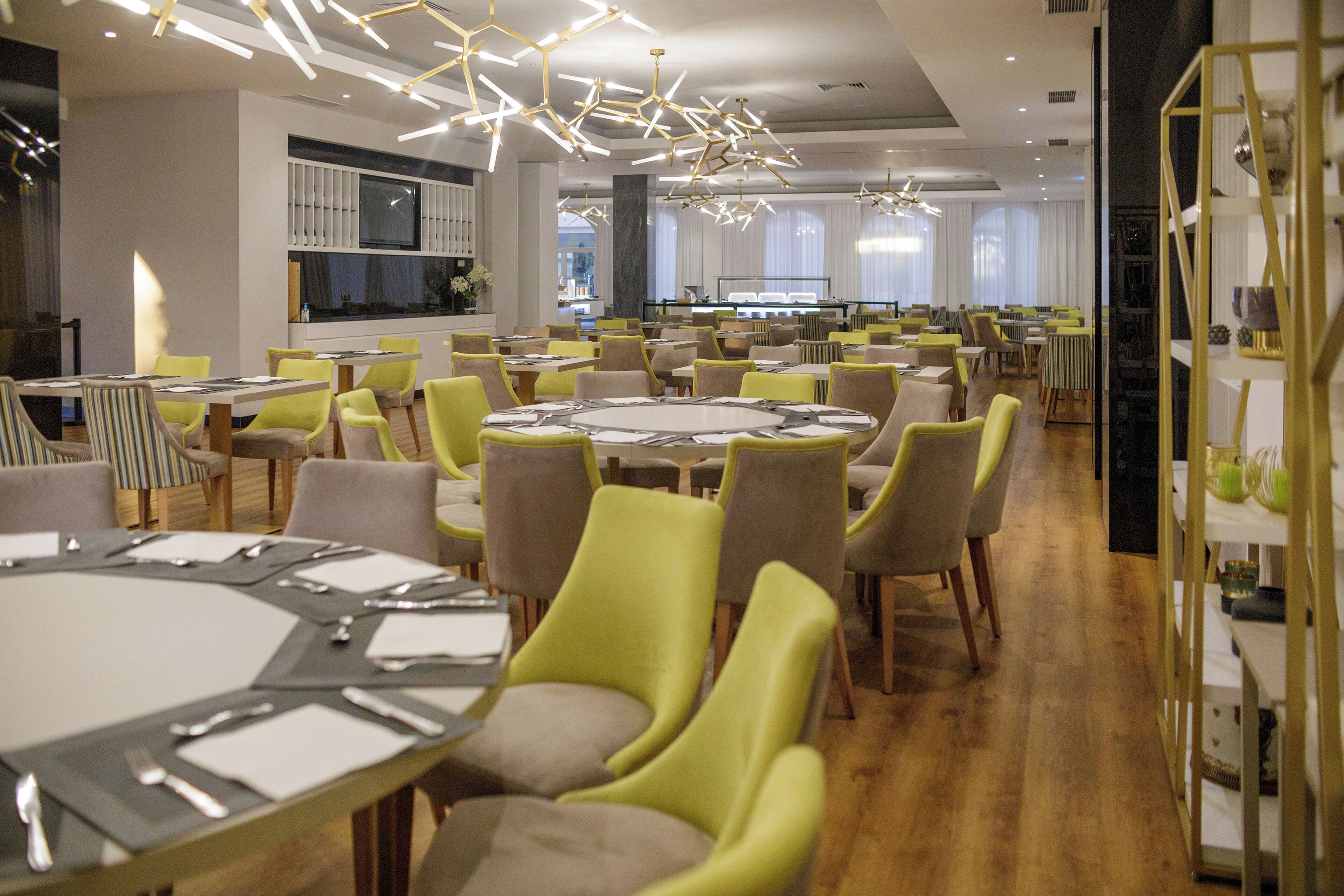 A modern dining area with multiple round tables and chairs, a large TV screen, and contemporary lighting fixtures.