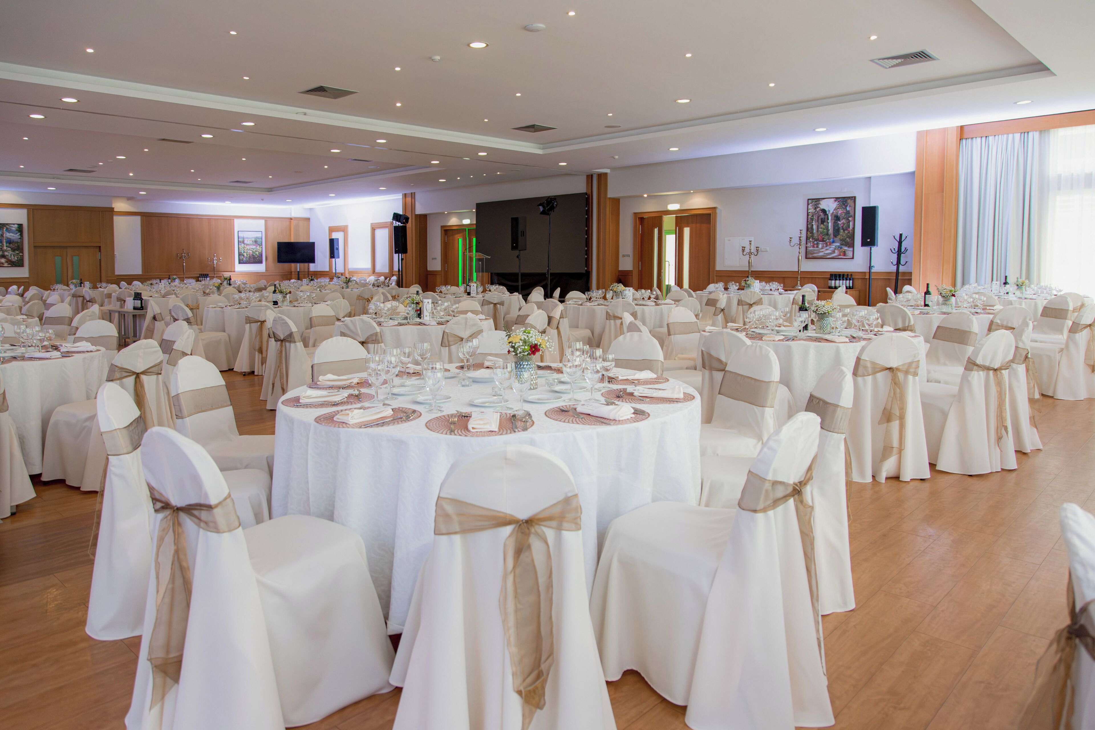 Un salón de banquetes con mesas redondas, sillas blancas con cintas doradas y un escenario con un orador y una pantalla.