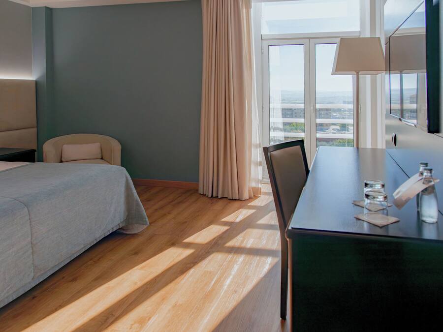 Quarto de hotel com cama, escrivaninha, cadeira, mesa de cabeceira, abajur, janela com cortinas e vista da cidade.
