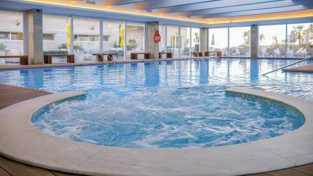 A large indoor swimming pool with a hot tub in the center, surrounded by lounge chairs and a wooden deck.
