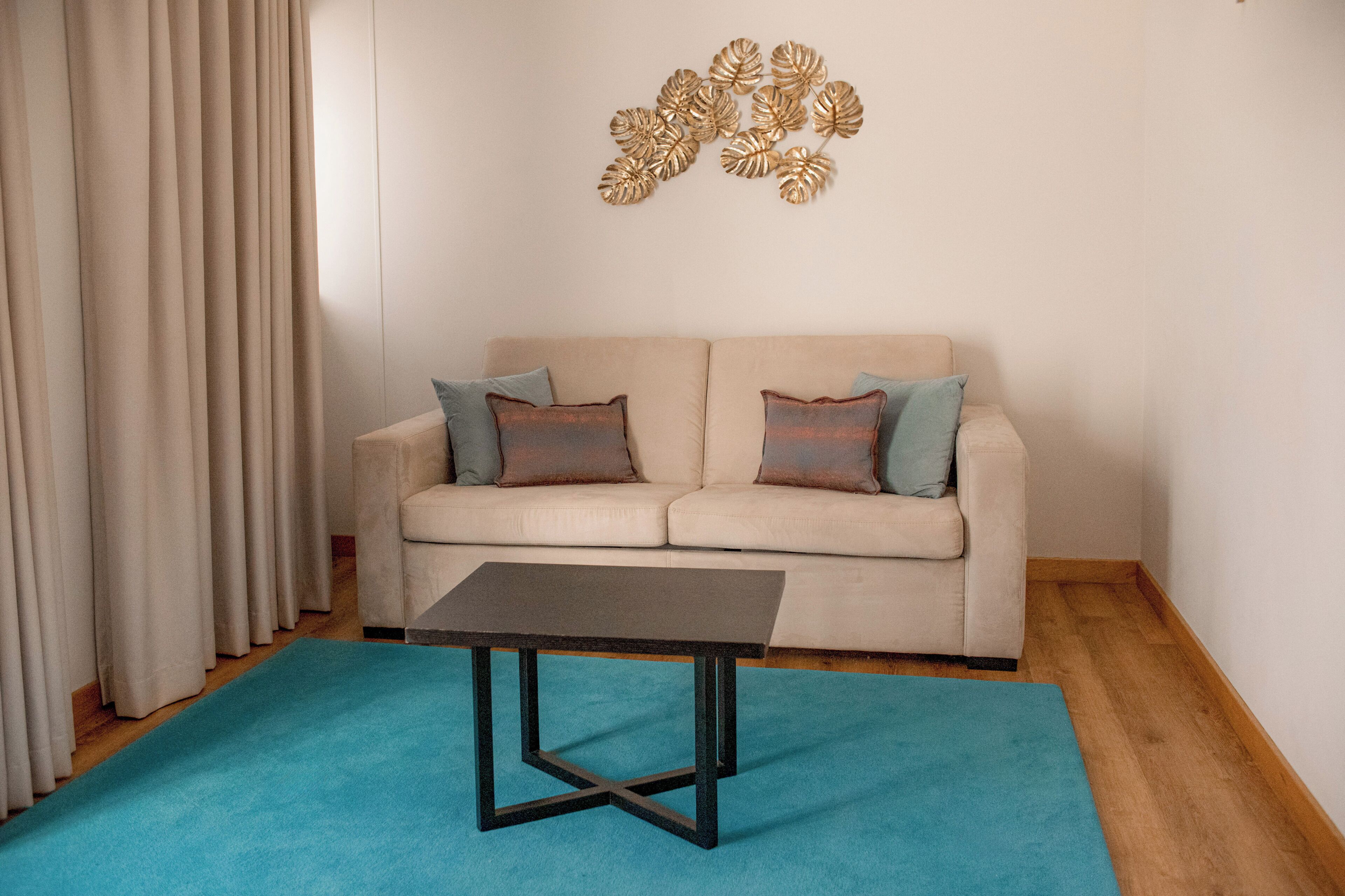 A living room with a beige sofa, two pillows, a blue rug, a small table, and a decorative wall piece.