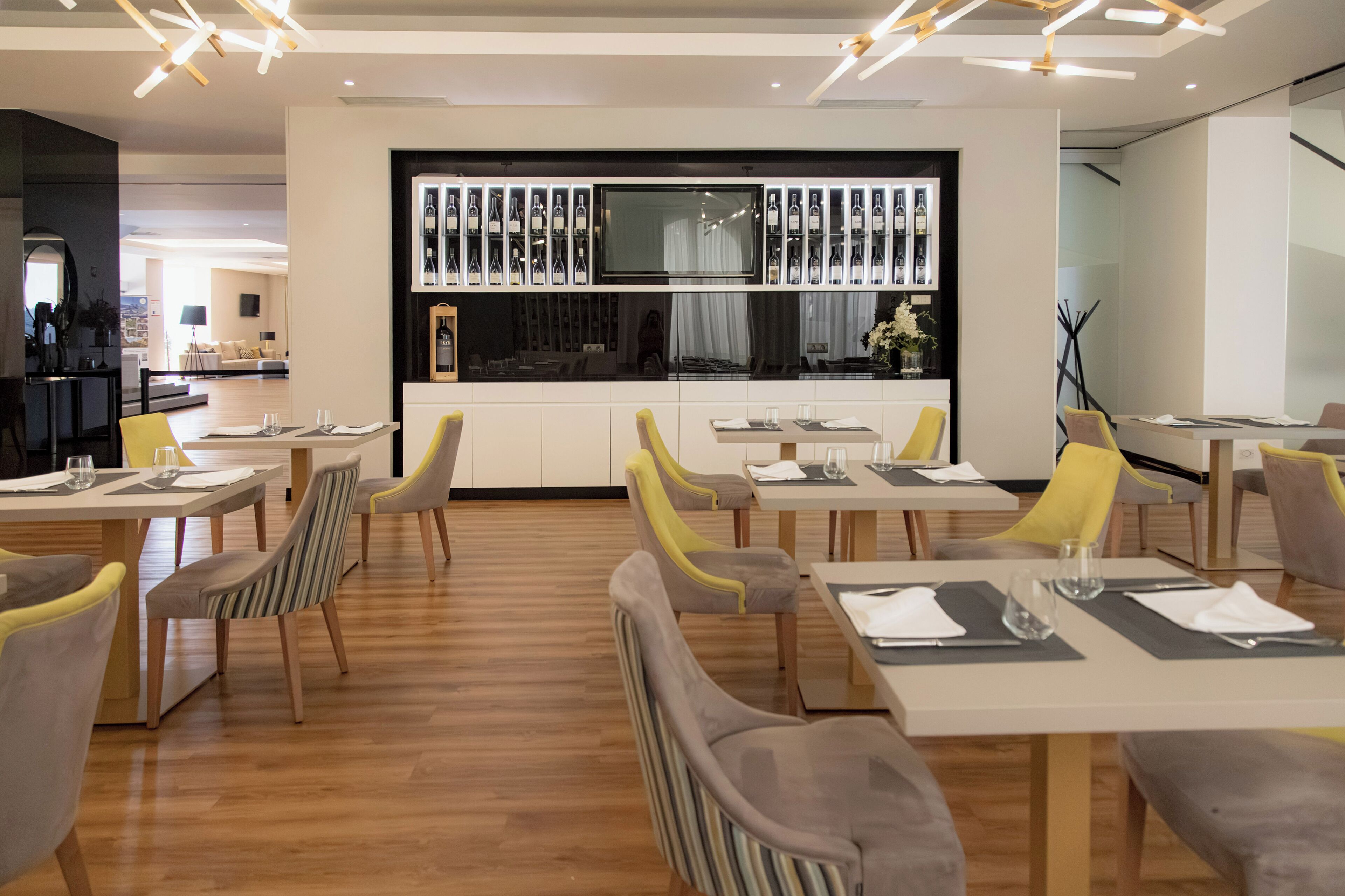 A modern dining area with wooden flooring, yellow and grey chairs, and a central TV screen displaying wine bottles.