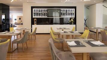 A modern dining area with wooden flooring, yellow and grey chairs, and a central TV screen displaying wine bottles.