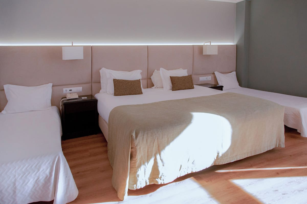 Habitación de hotel con dos camas individuales, una cama más grande, una mesita de noche y una lámpara fijada en la pared.