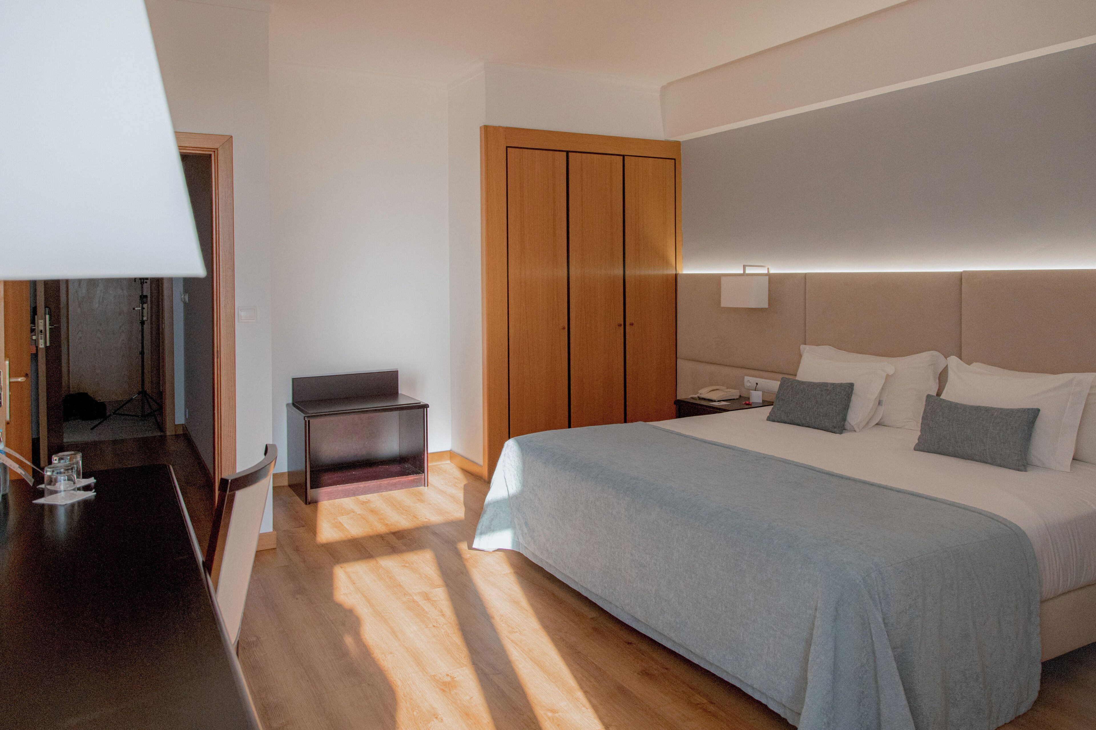 A modern hotel room with a large bed, a wooden wardrobe, a desk, and a bedside table.