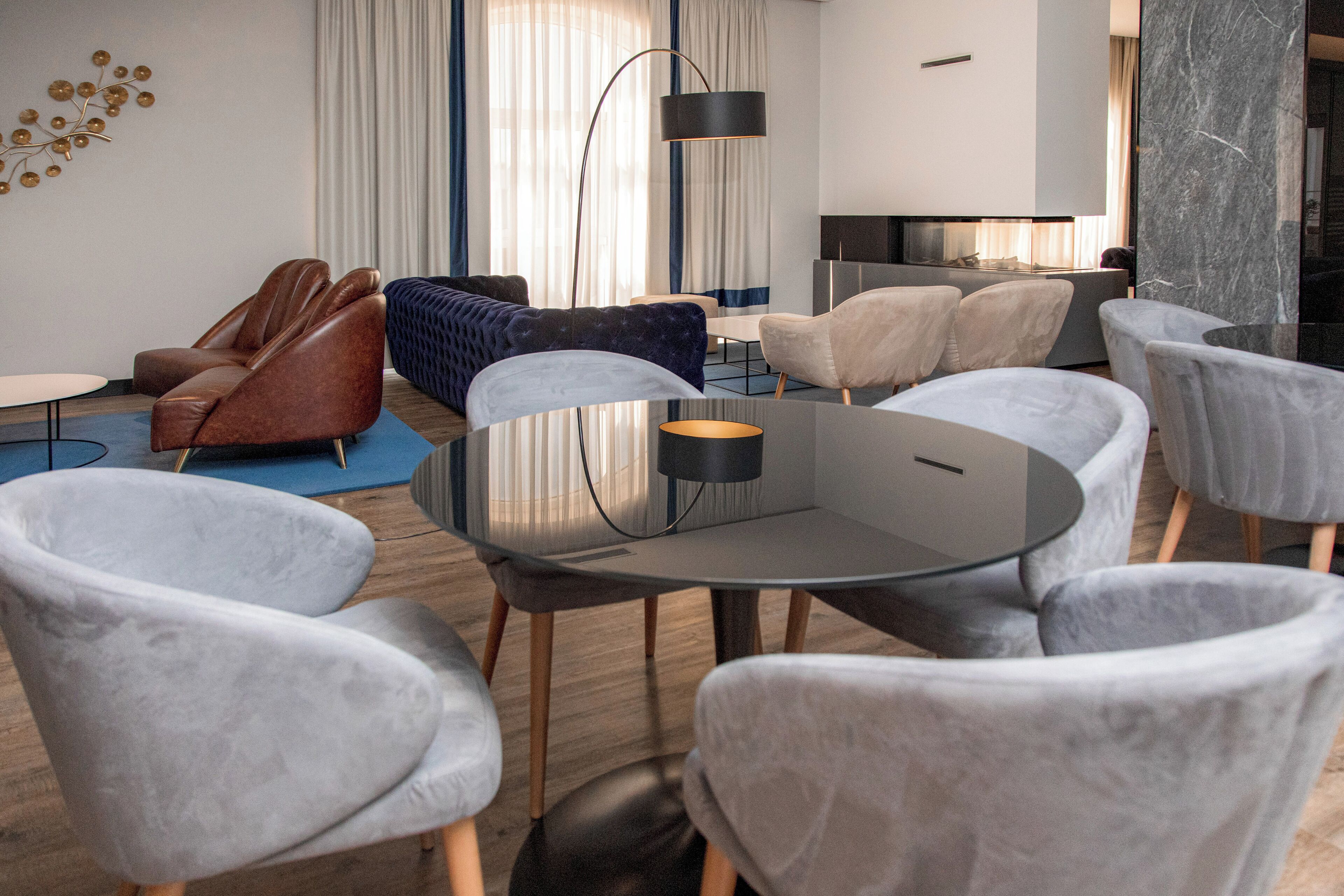 Una sala de estar moderna con una mesa redonda de cristal, sillas tapizadas en gris, un sofá de cuero marrón y un tapete azul estampado.