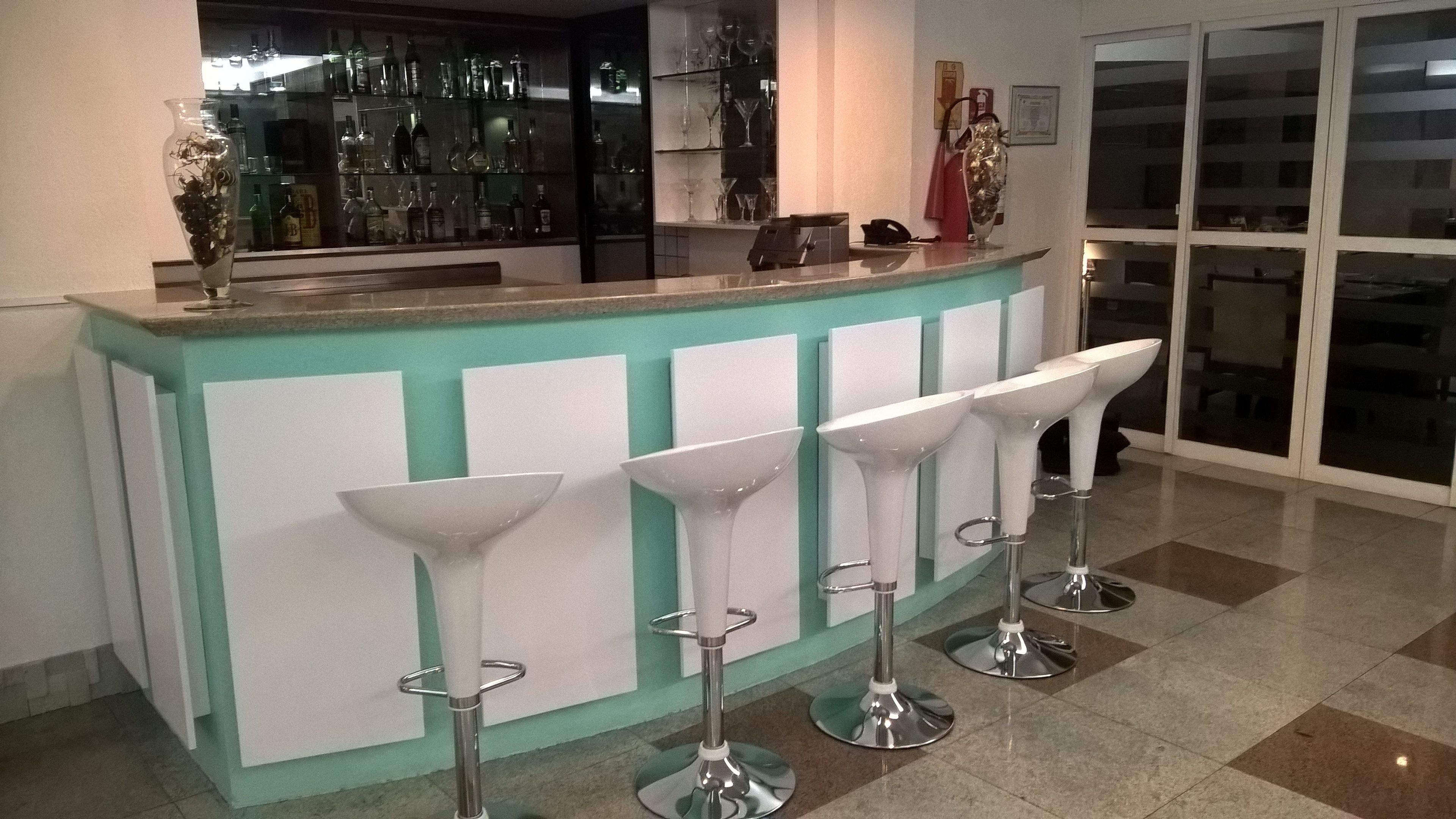 Un espace bar doté d’un comptoir turquoise, de quatre tabourets de bar blancs et d’une vitrine en verre où sont exposées des bouteilles.