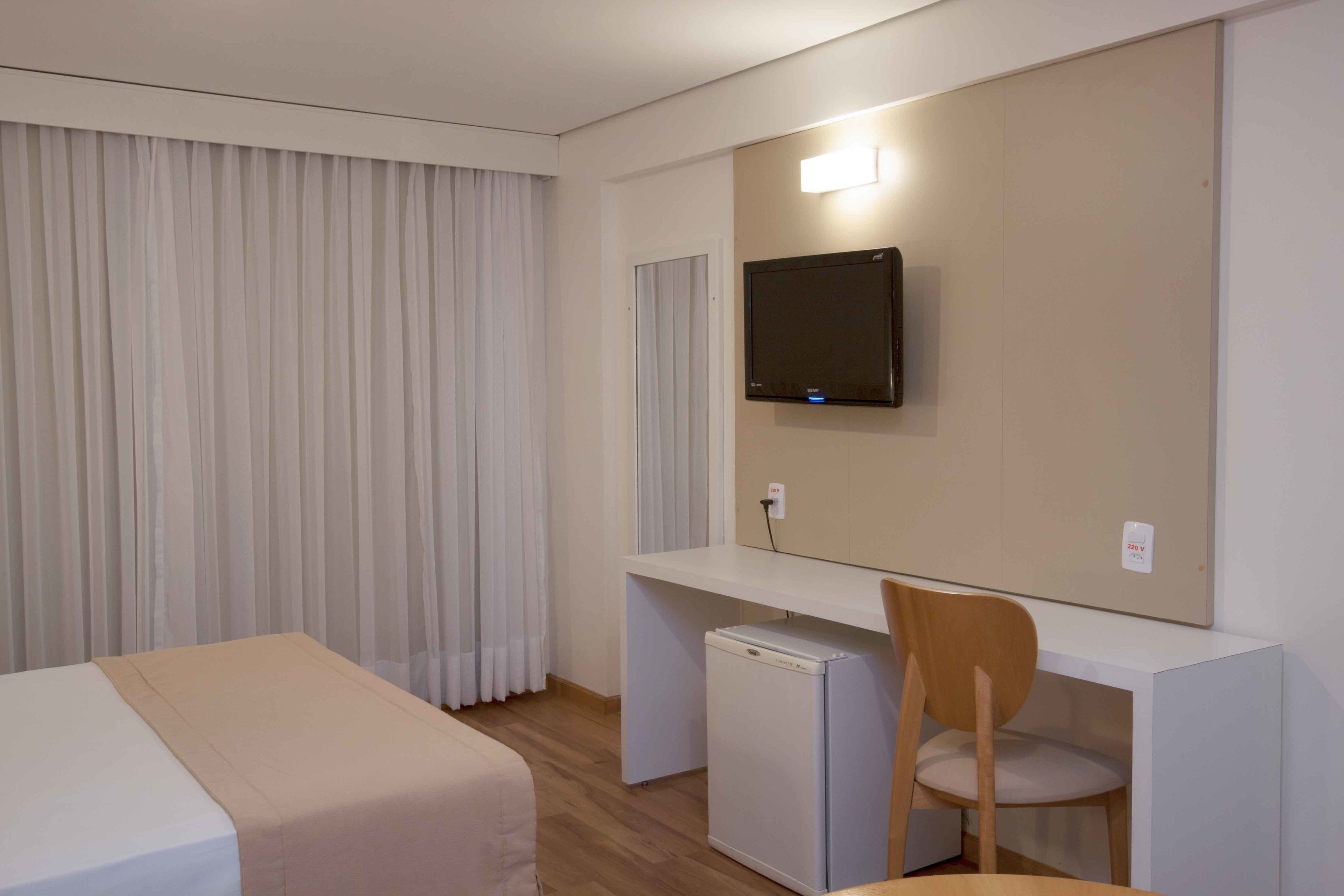 Habitación de hotel con cama, escritorio, silla, televisor y refrigerador pequeño.