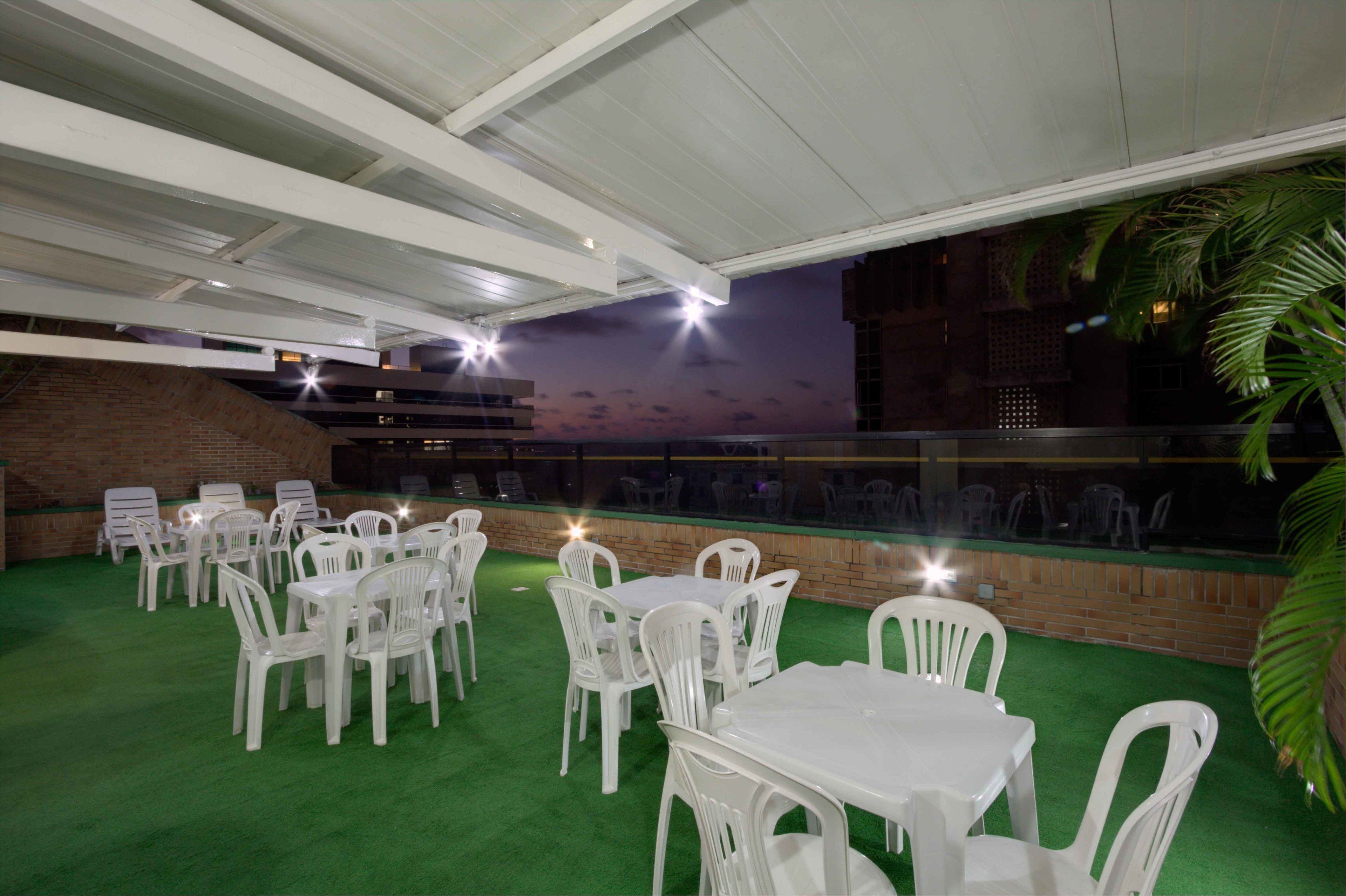 Un espacio de comedor al aire libre con sillas y mesas de plástico blanco, un muro de ladrillo y vista al atardecer.