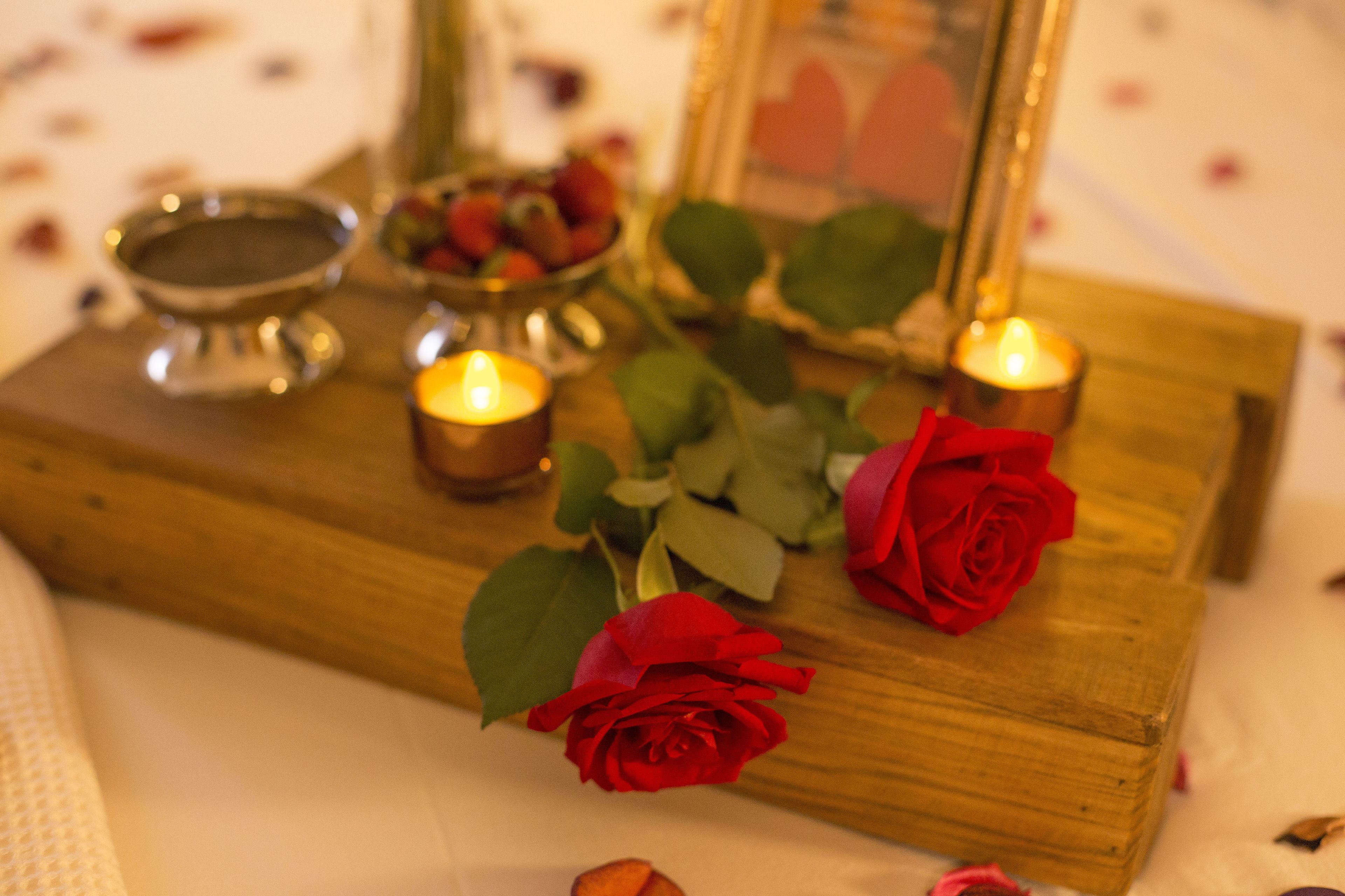 A wooden table with candles, a bowl of strawberries, and a red rose.