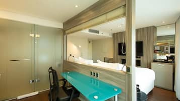 Una habitación de hotel moderna con una cama grande, un escritorio con silla, una kitchenette pequeña y un baño con ducha e inodoro.