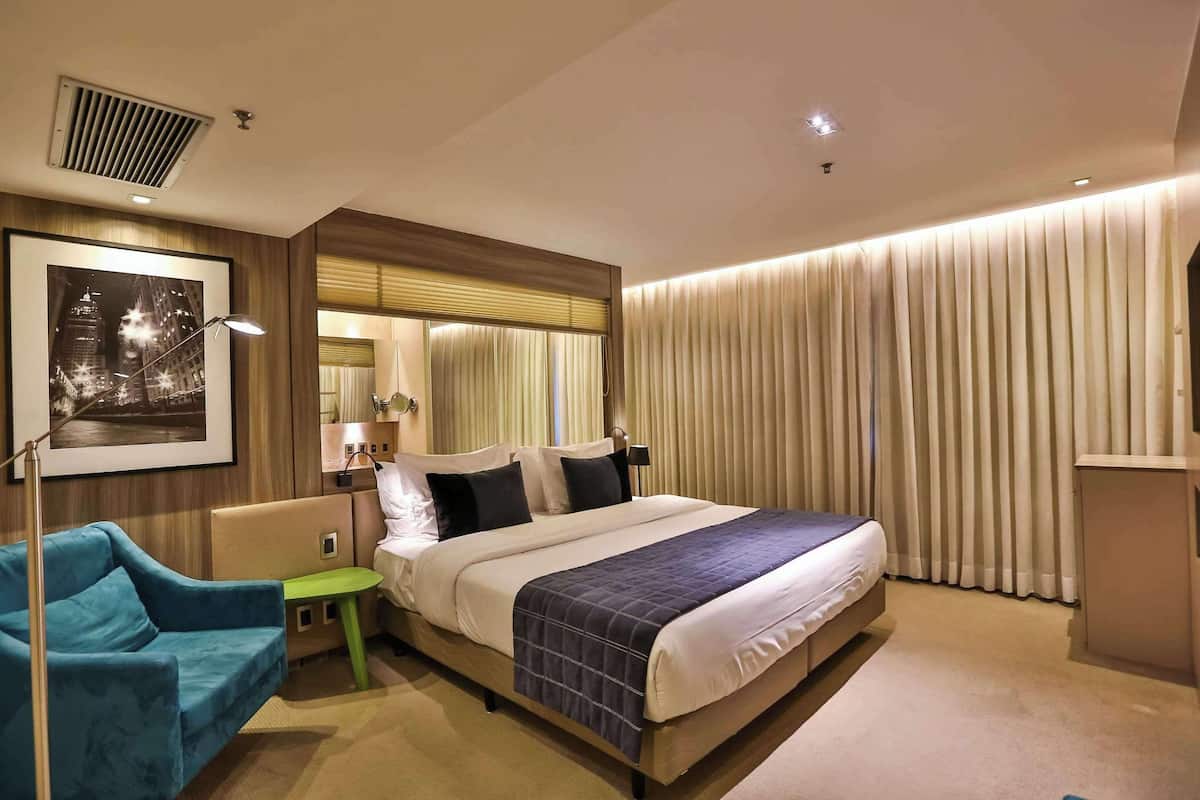 Une chambre d’hôtel avec un grand lit, un bureau, une chaise, un tableau encadré au mur et une fenêtre avec des rideaux.
