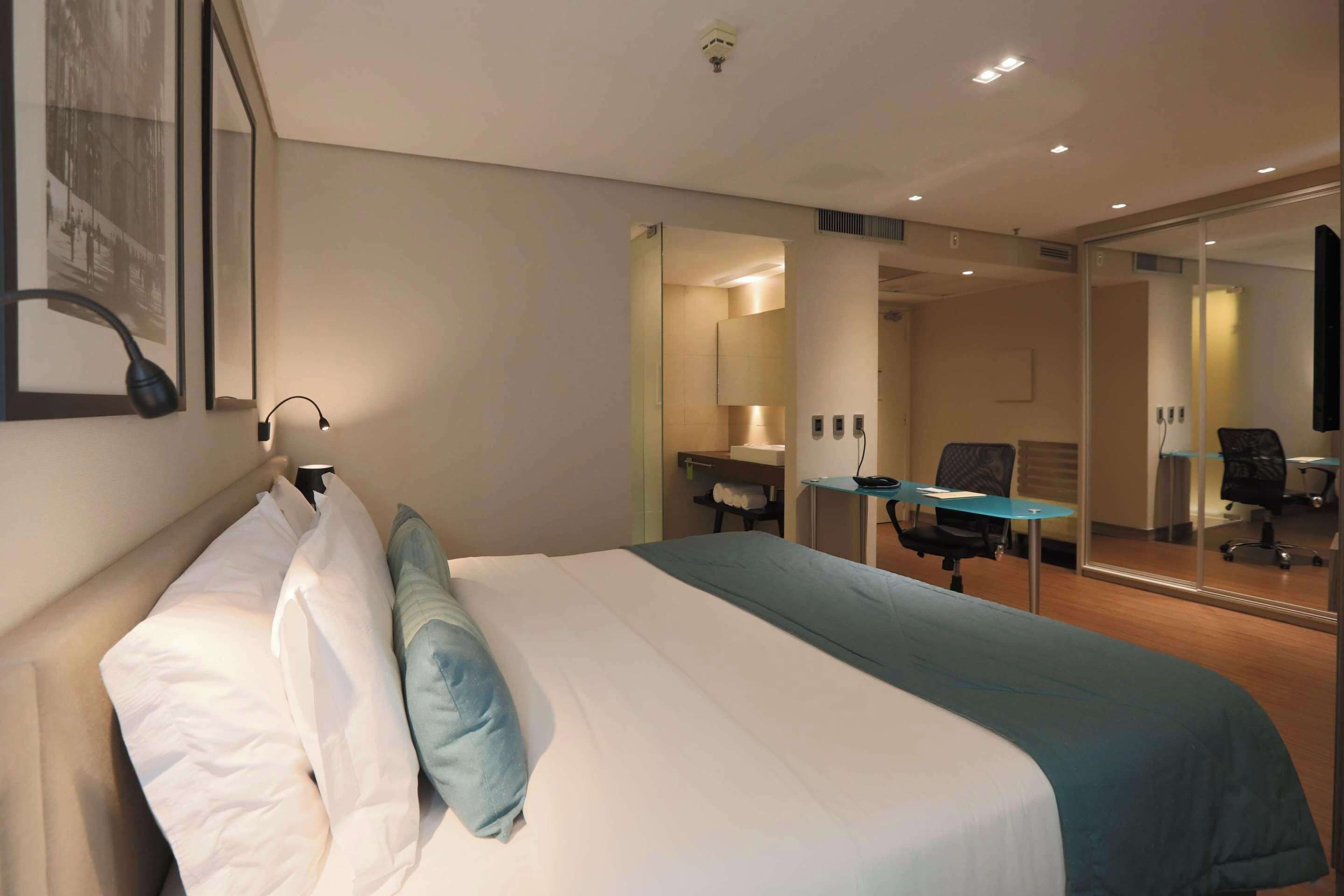 A hotel room with a large bed, a desk, and a chair. There is a mirror on the wall and a window with a picture frame.