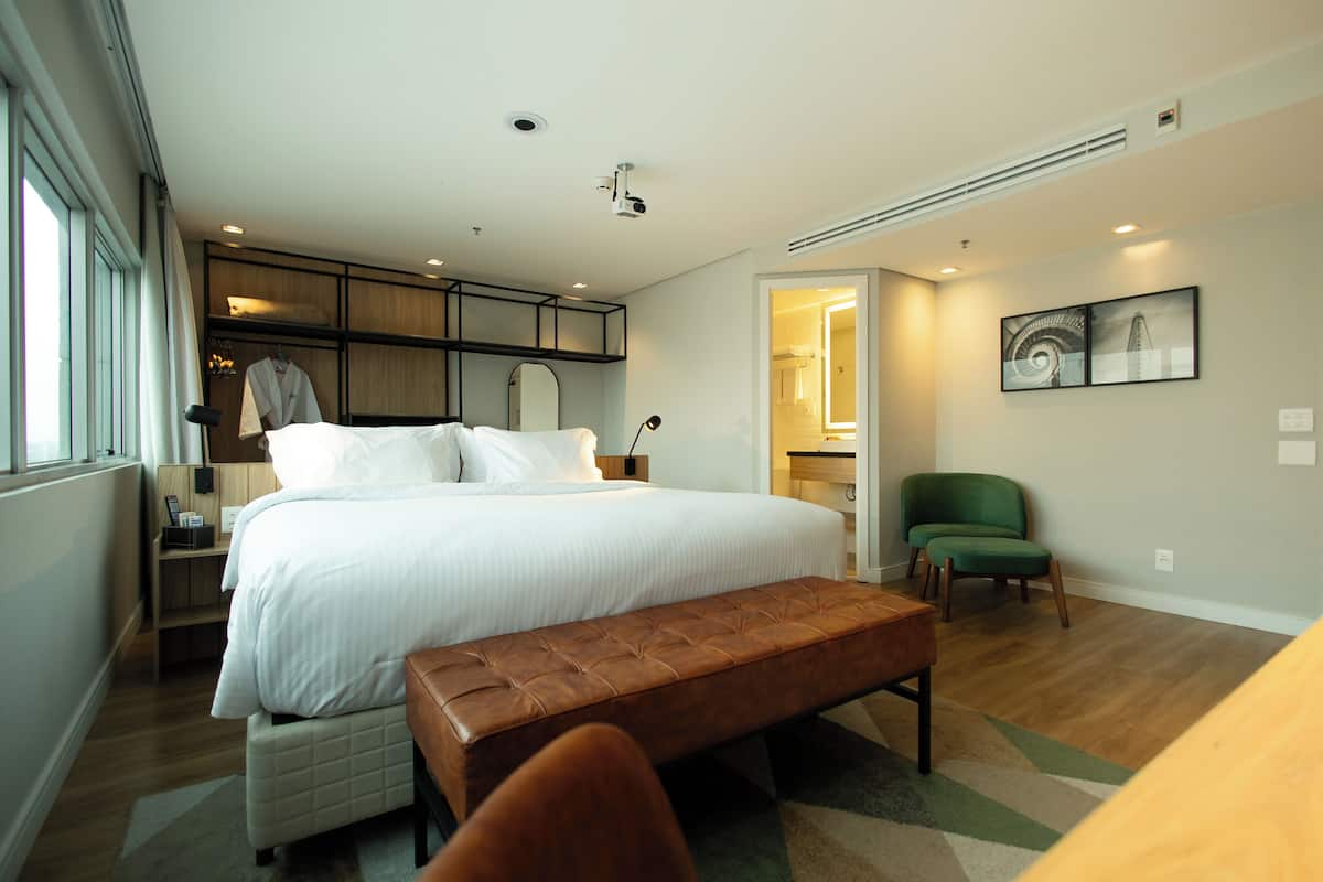 Une chambre d’hôtel moderne avec un grand lit, un banc, un fauteuil vert et une vue sur la ville par la fenêtre.