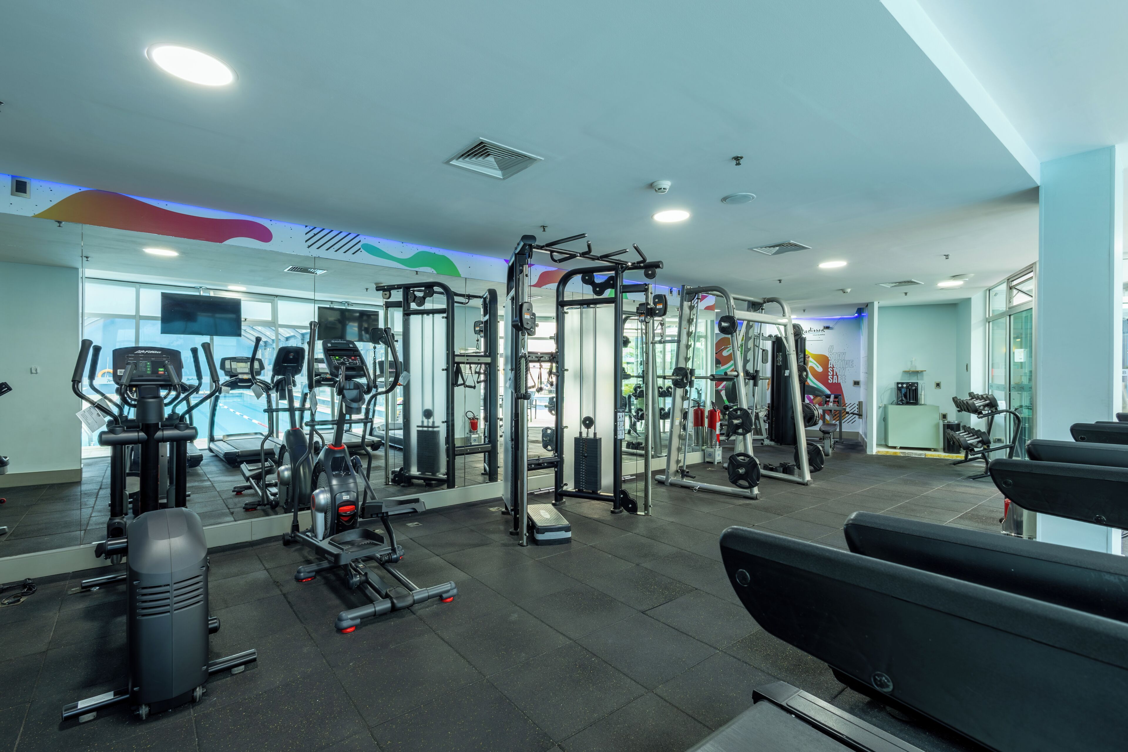 Un gimnasio con diferentes equipos de ejercicio, espejos y un ventanal grande.