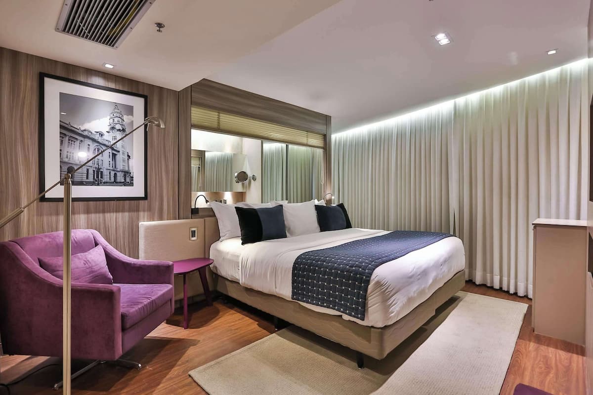 Une chambre d’hôtel moderne avec un grand lit, un fauteuil violet, un bureau et un tableau encadré au mur.