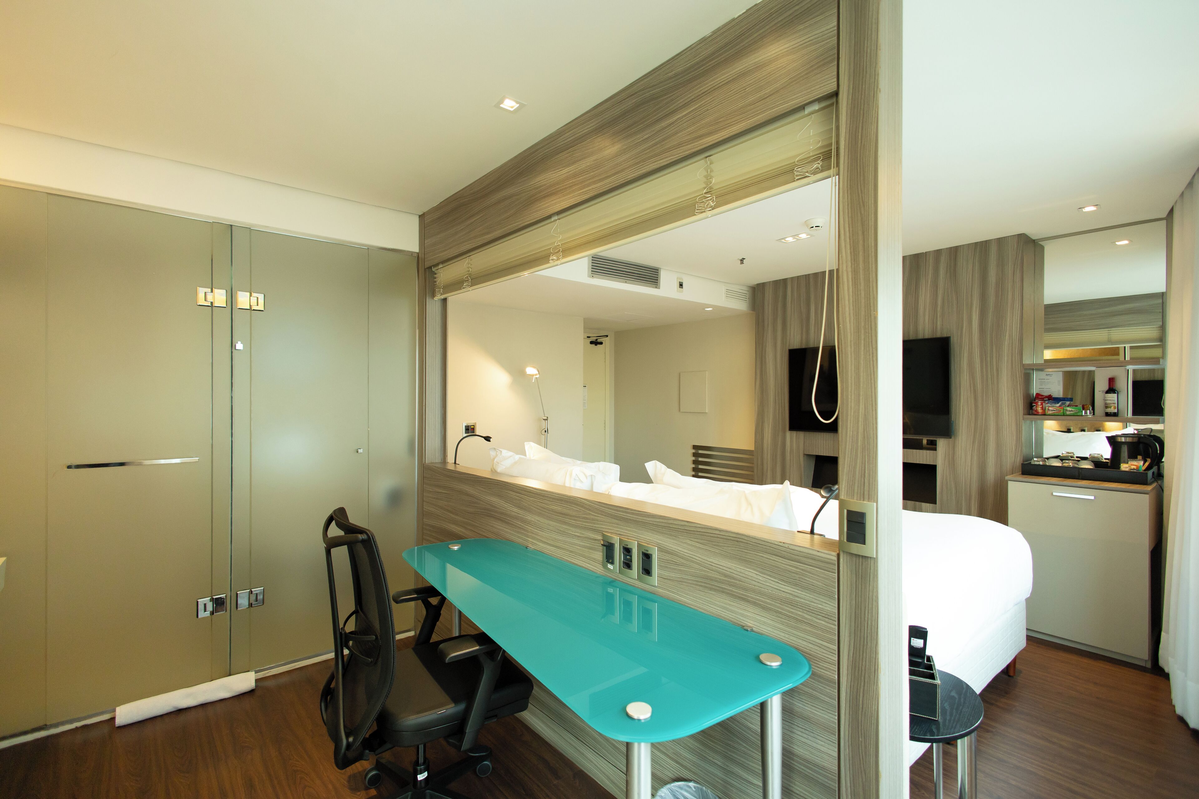 A modern hotel room with a bed, a desk, a chair, a mirror, and a kitchenette.