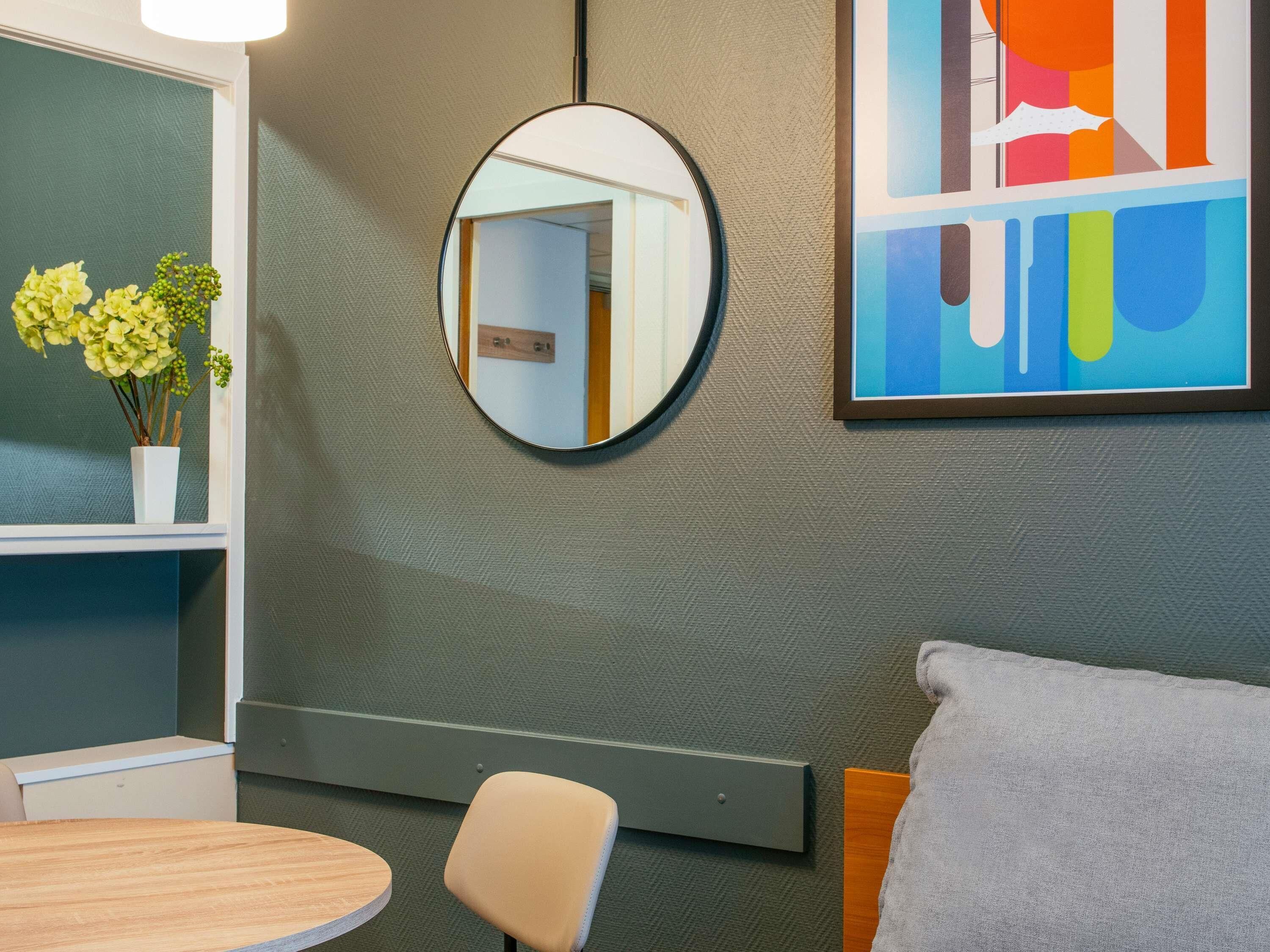 A modern bathroom with a round mirror, a wooden chair, a small table, and a colorful abstract painting on the wall.
