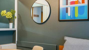 A modern bathroom with a round mirror, a wooden chair, a small table, and a colorful abstract painting on the wall.