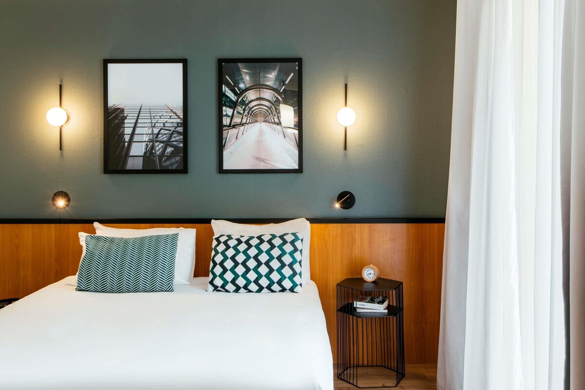 A hotel room with a bed, two wall-mounted lights, two framed pictures, a bedside table with a clock, and a curtain.